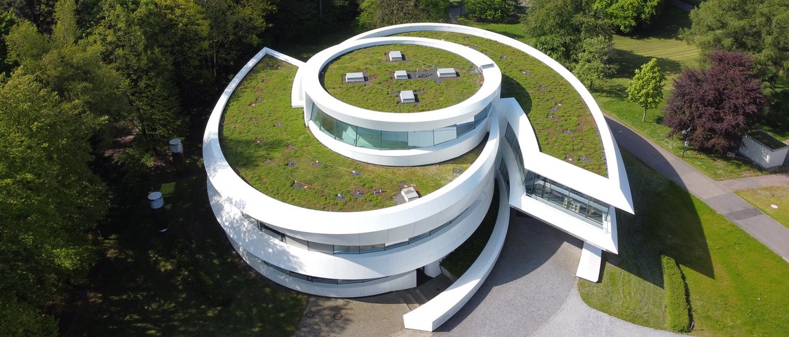 Drohnenaufnahme des HdA-Gebäudes mit begrüntem Dach von oben eingeräahmt von Bäumen, im Hintergrund die Teleskopkuppeln des Elsässer-Labors