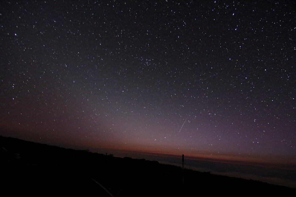 Picture of the night sky underlaid by a faint, oblique wedge-shaped glow and a shooting star