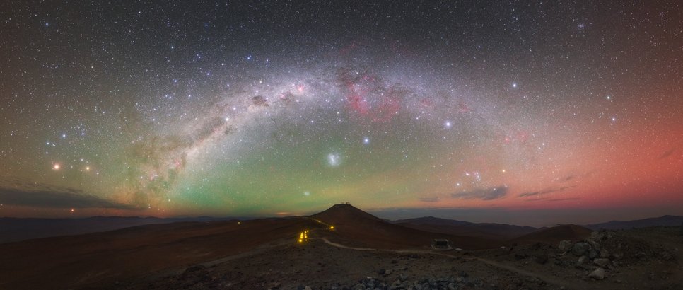 Panoramic view of the Milky Way as an arc over Paranal. The night sky is brightened by a green glow in the center and in red to the right edge
