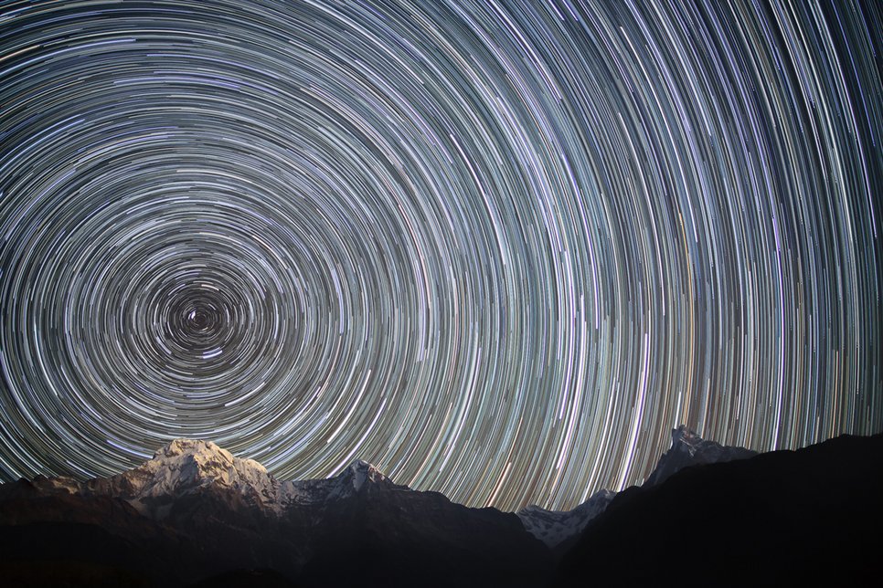 Star trails of many stars, whose circle segments form circles around the northern celestial pole. Below a snowy mountain landscape