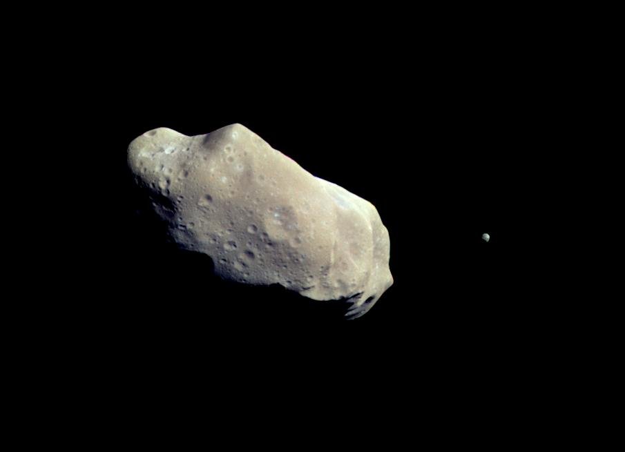 A potato-shaped, gray-brown object with a cratered surface and a small moon on a black background