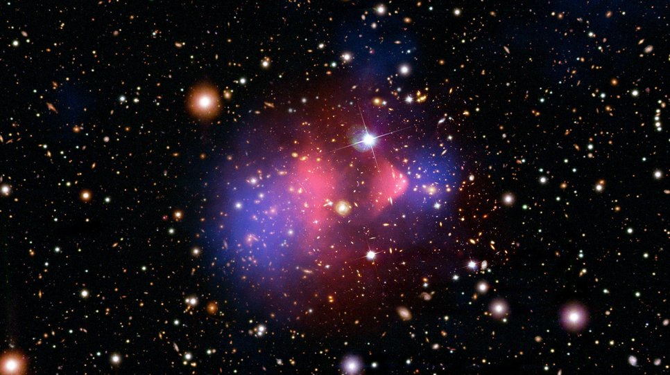 The Bullet Cluster: Hubble image of a galaxy cluster overlaid with two false-color images in pink and blue, marking a concentration in the center with a wedge-shaped compression and two separate regions outside